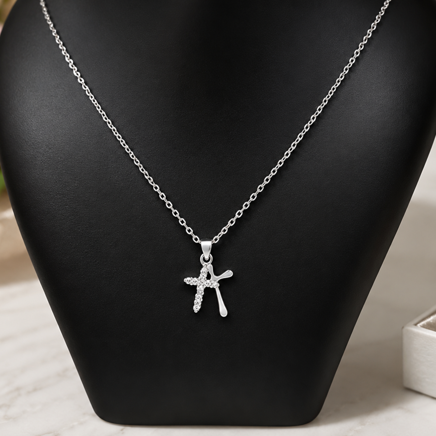 A silver cross pendant necklace with small clear stones, displayed on a black jewelry bust against a softly blurred light background with pink flowers and a jewelry box.