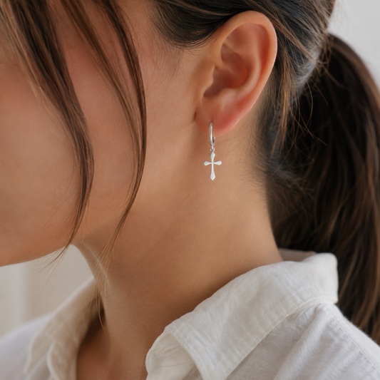 A close-up of a woman's ear wearing a delicate silver cross dangle earring, with her hair tied back and a white collared shirt visible.