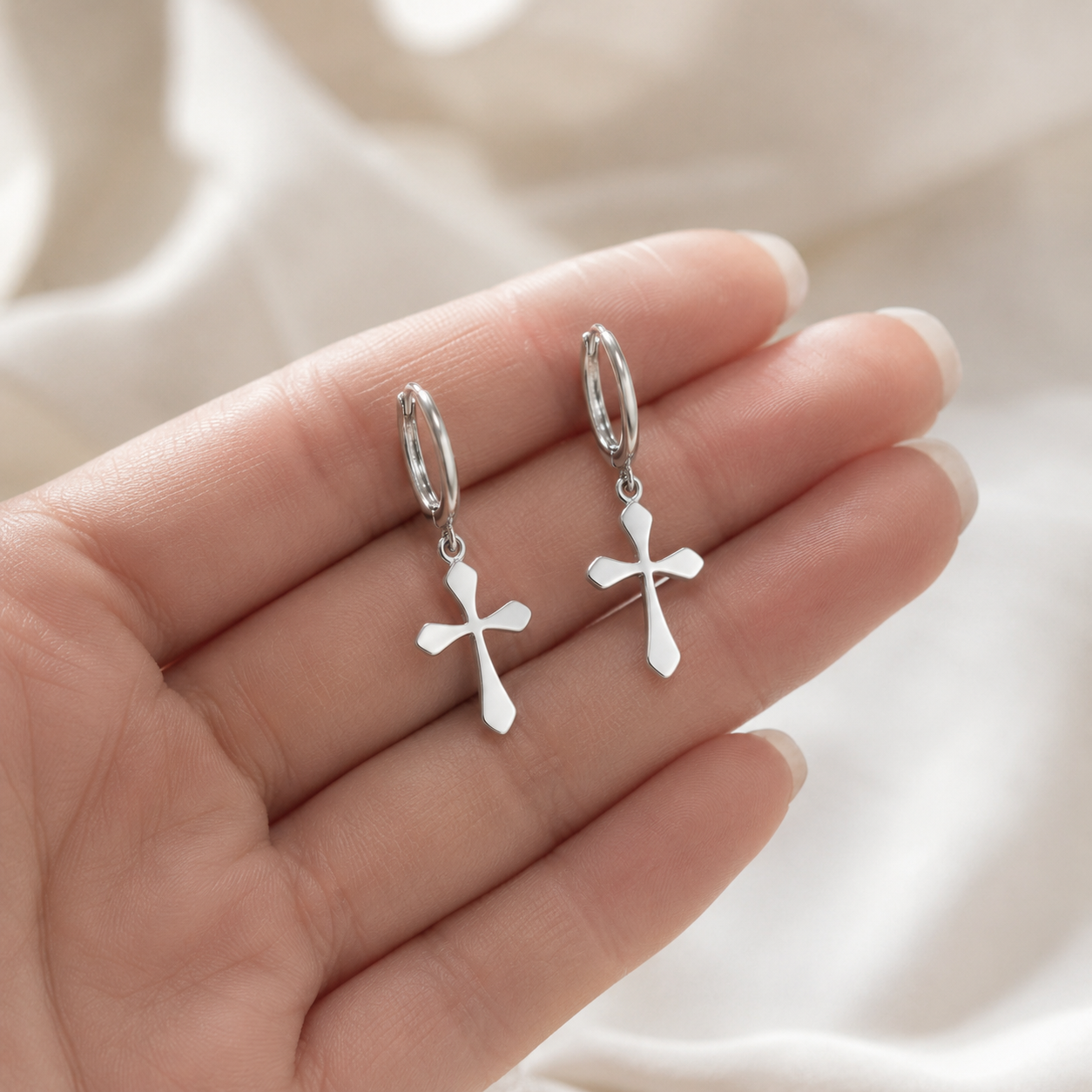 A pair of silver cross dangle earrings held in a person's hand against a soft white fabric background.