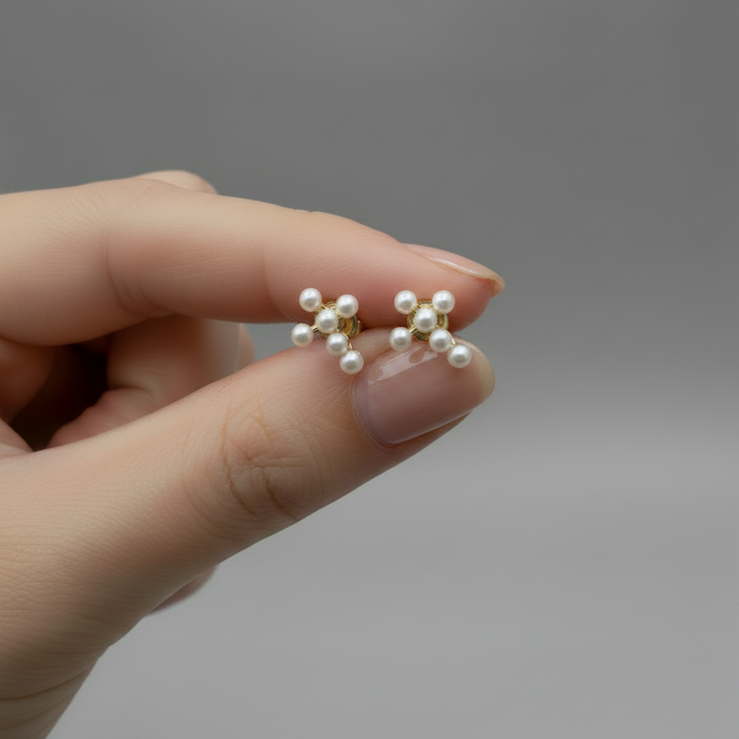A close-up of a hand holding a pair of small gold stud earrings with clustered white pearls against a soft gray background.