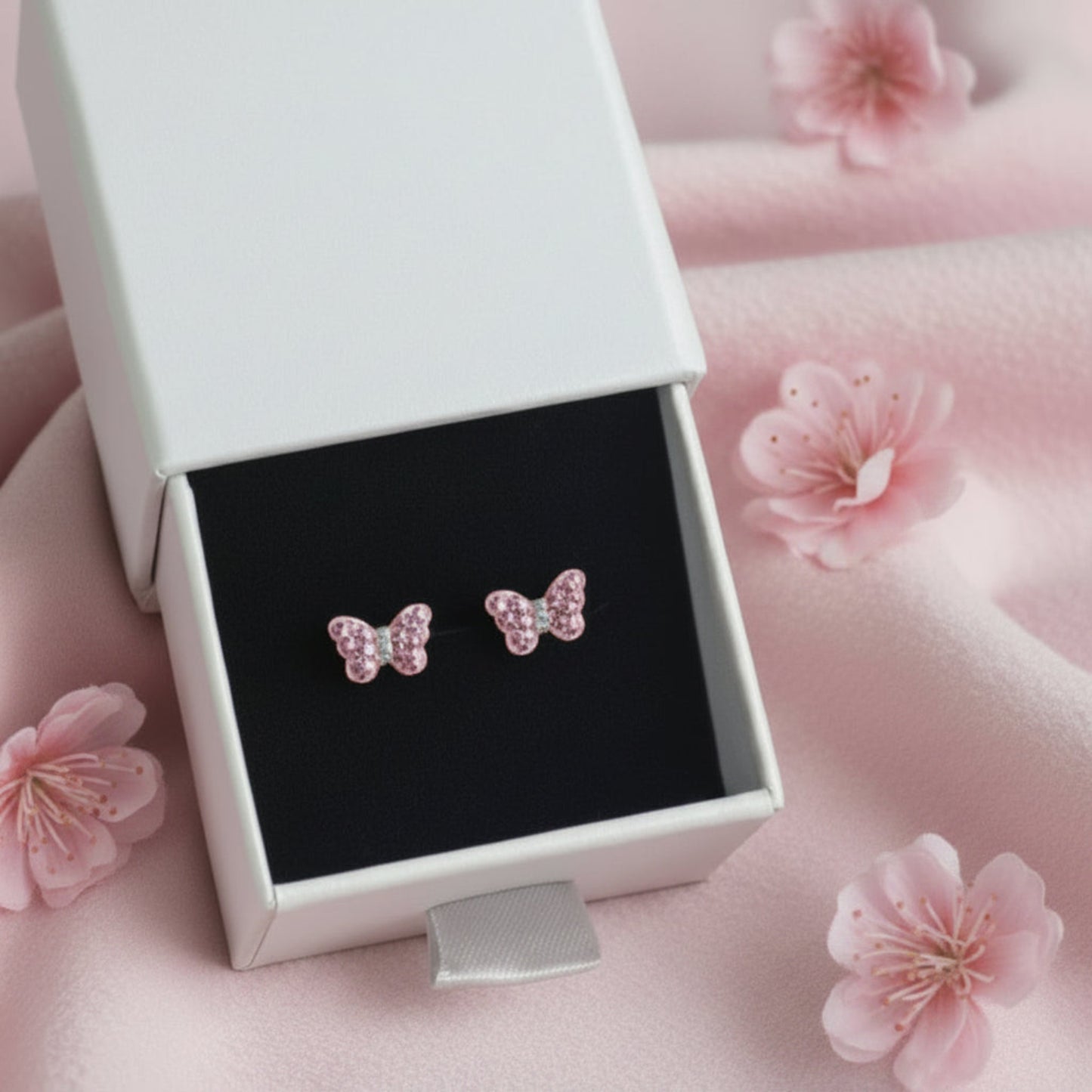 A pair of small butterfly-shaped stud earrings with pink rhinestones, displayed in an open light grey jewellery box with a black insert on a pink surface.