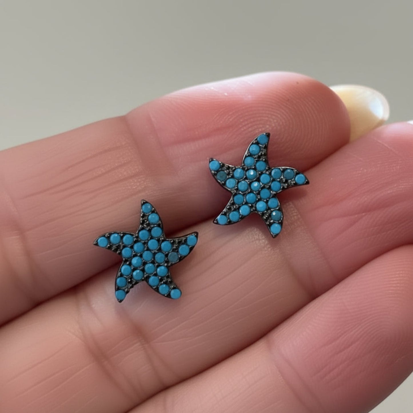 A close-up of a person's hand holding a pair of starfish-shaped stud earrings covered in small blue rhinestones.
