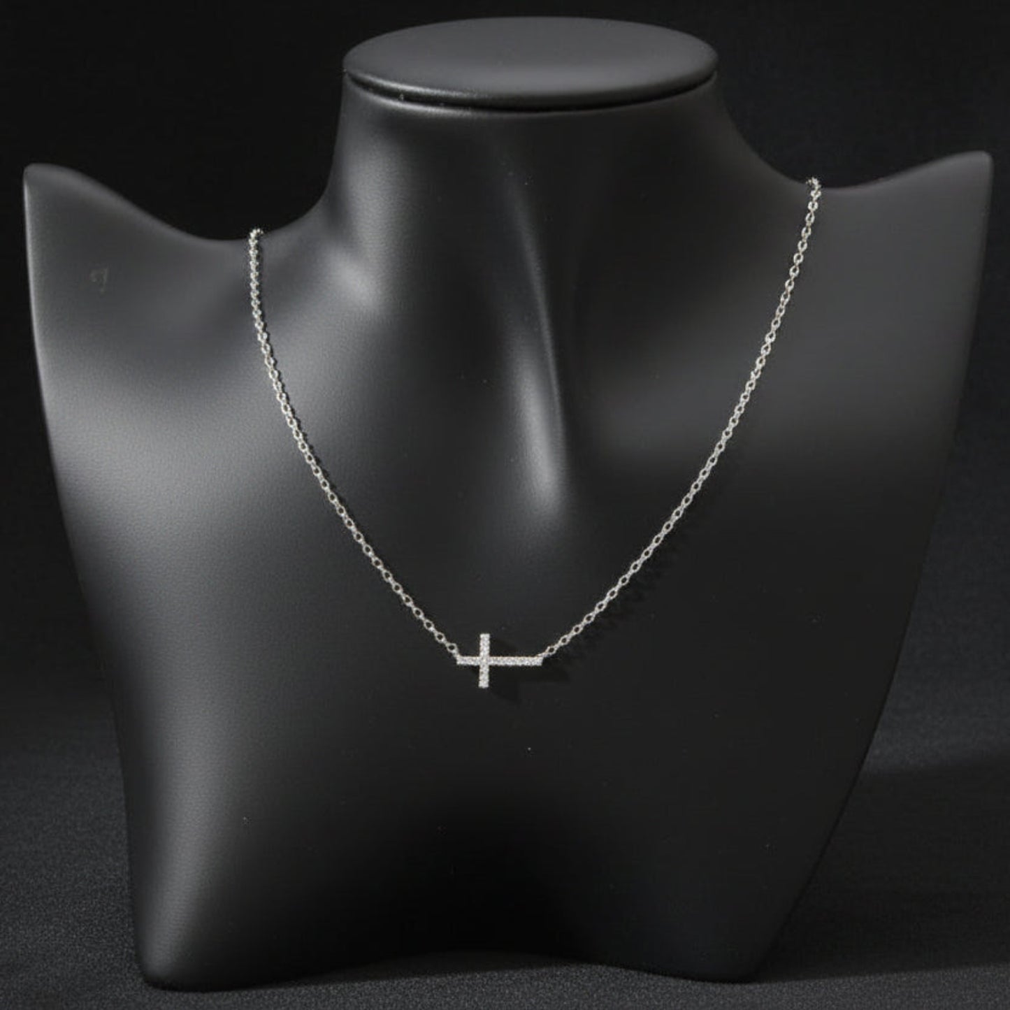 A delicate silver cross pendant necklace displayed on a matte black jewelry bust against a light gray background.