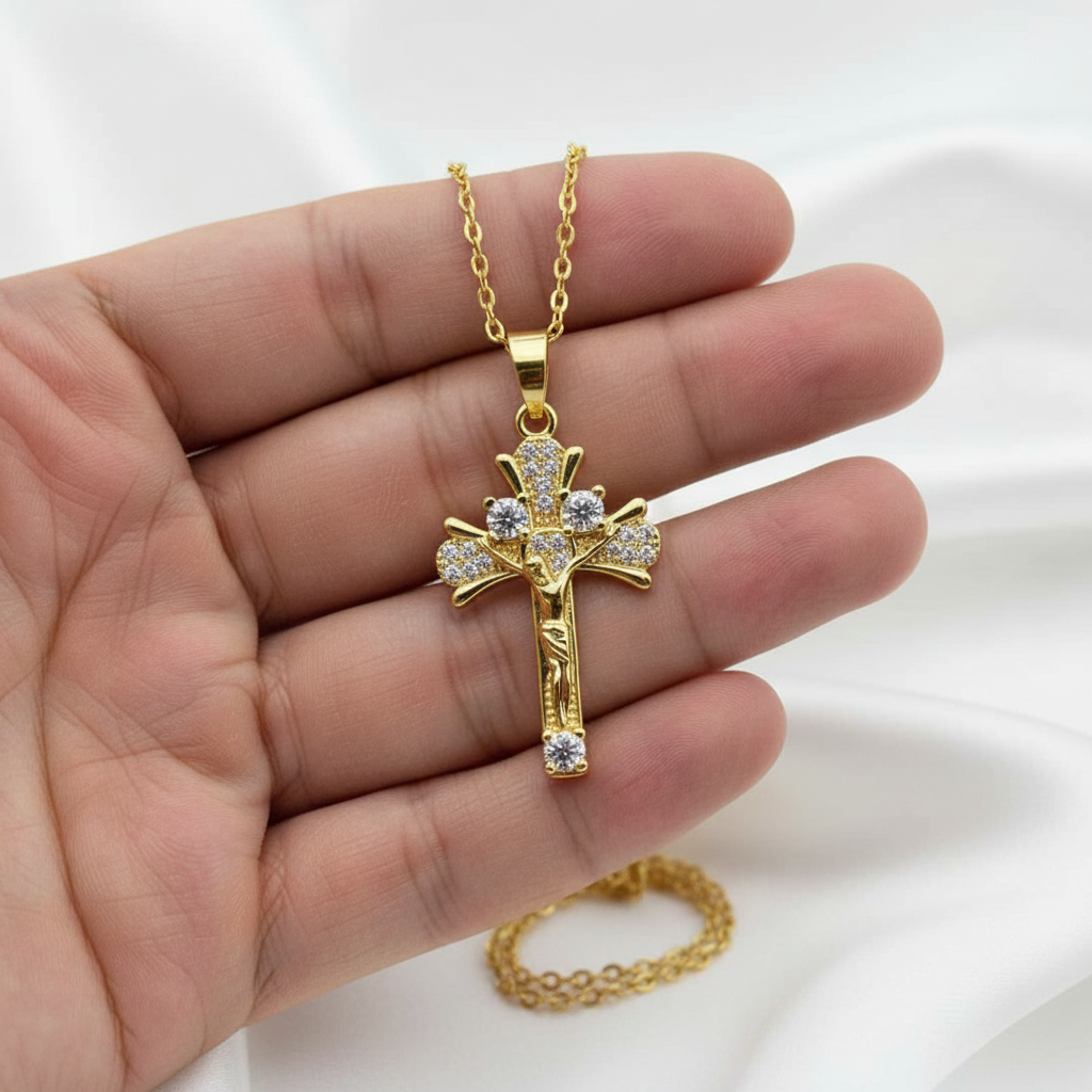 A gold cross pendant with clear stones, displayed on a person's hand against a white fabric background.