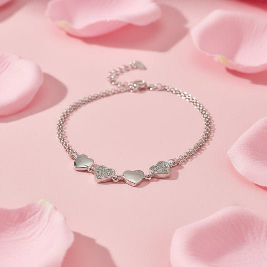 A silver chain bracelet with three heart charms, including two small heart charms covered in clear pavé stones, displayed on a white surface.