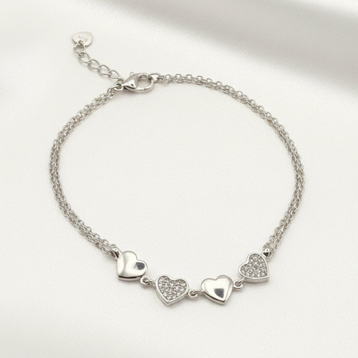 A silver chain bracelet with three dangling heart charms, displayed on a white satin fabric background.