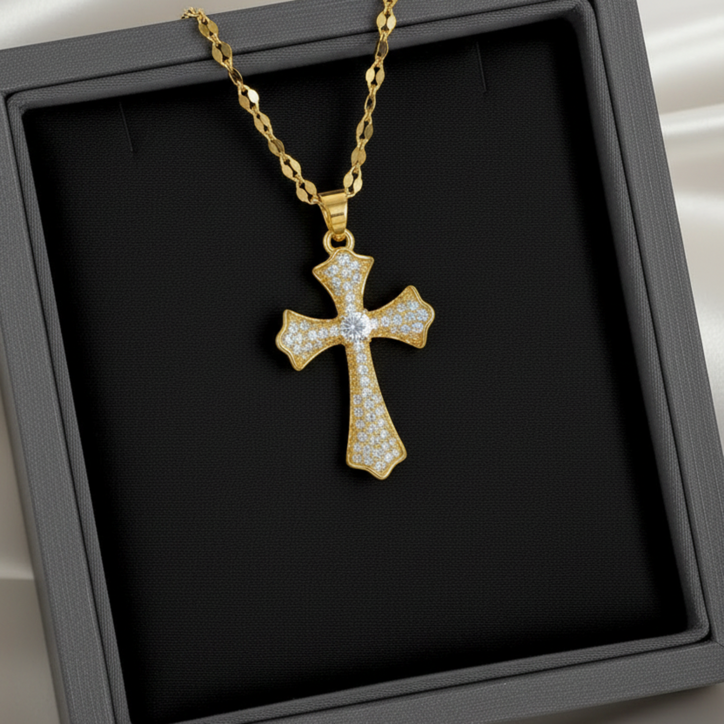A gold cross pendant necklace paved with small clear stones, displayed on a black jewelry box with a white satin background.