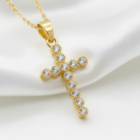 A gold cross pendant necklace adorned with small clear gemstones, resting on draped white satin fabric.