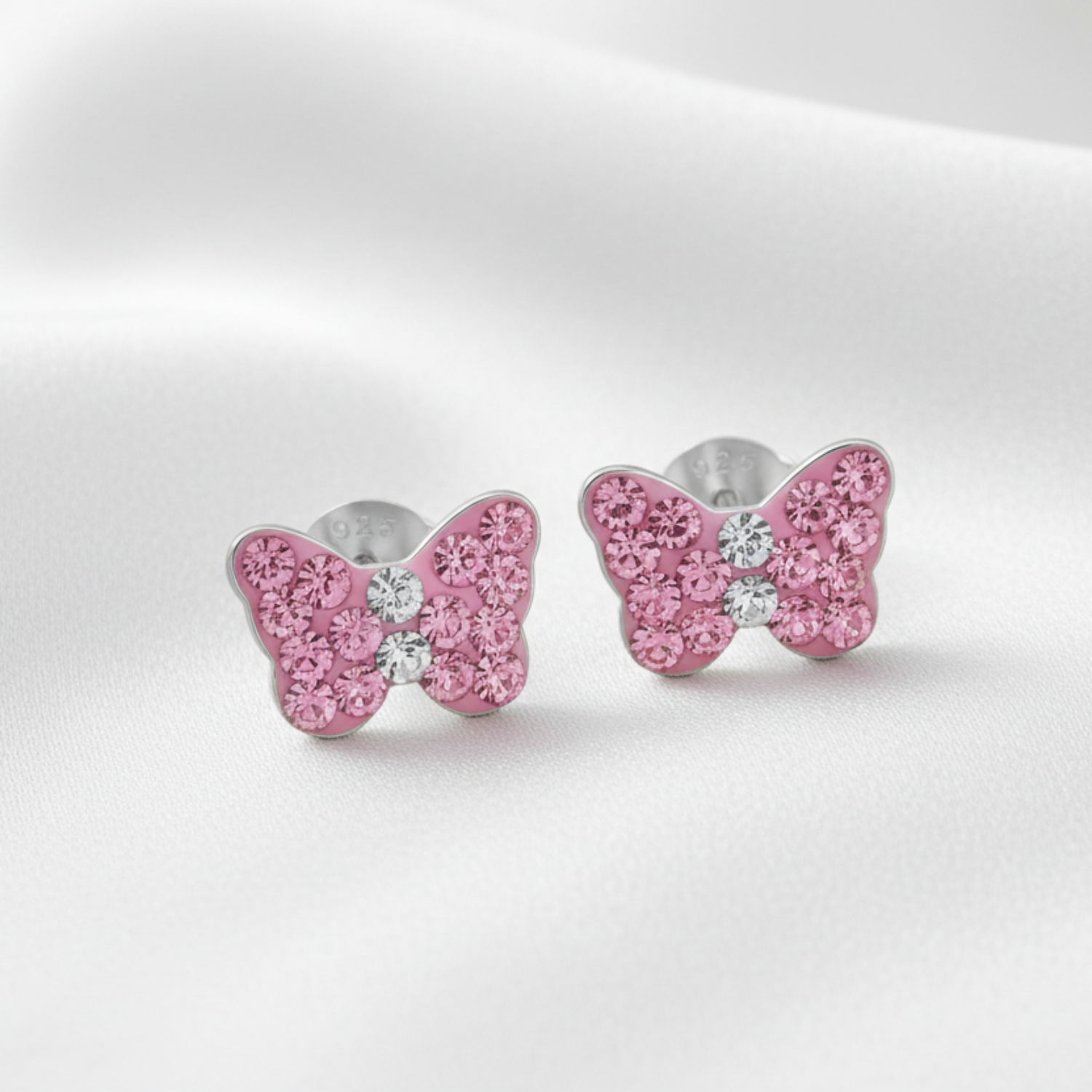 A pair of butterfly-shaped stud earrings with sparkling pink stones and a small clear center stone, set in silver-toned metal on a soft white fabric background.