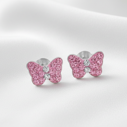 A pair of butterfly-shaped stud earrings with sparkling pink stones and a small clear center stone, set in silver-toned metal on a soft white fabric background.