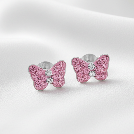A pair of butterfly-shaped stud earrings with sparkling pink stones and a small clear center stone, set in silver-toned metal on a soft white fabric background.