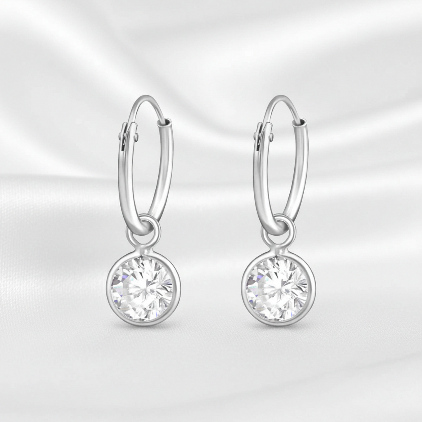A pair of silver hoop earrings with round bezel-set clear gemstones, displayed on a white satin fabric background.