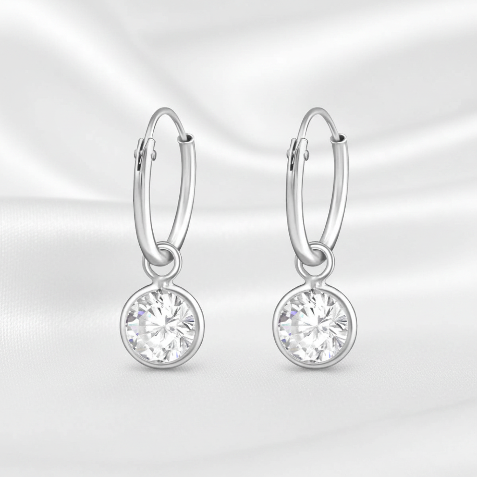 A pair of silver hoop earrings with round bezel-set clear gemstones, displayed on a white satin fabric background.