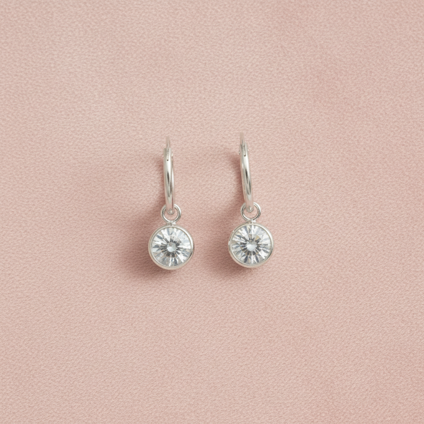 A pair of silver dangle earrings with round bezel-set clear crystals, displayed on a textured light pink background.