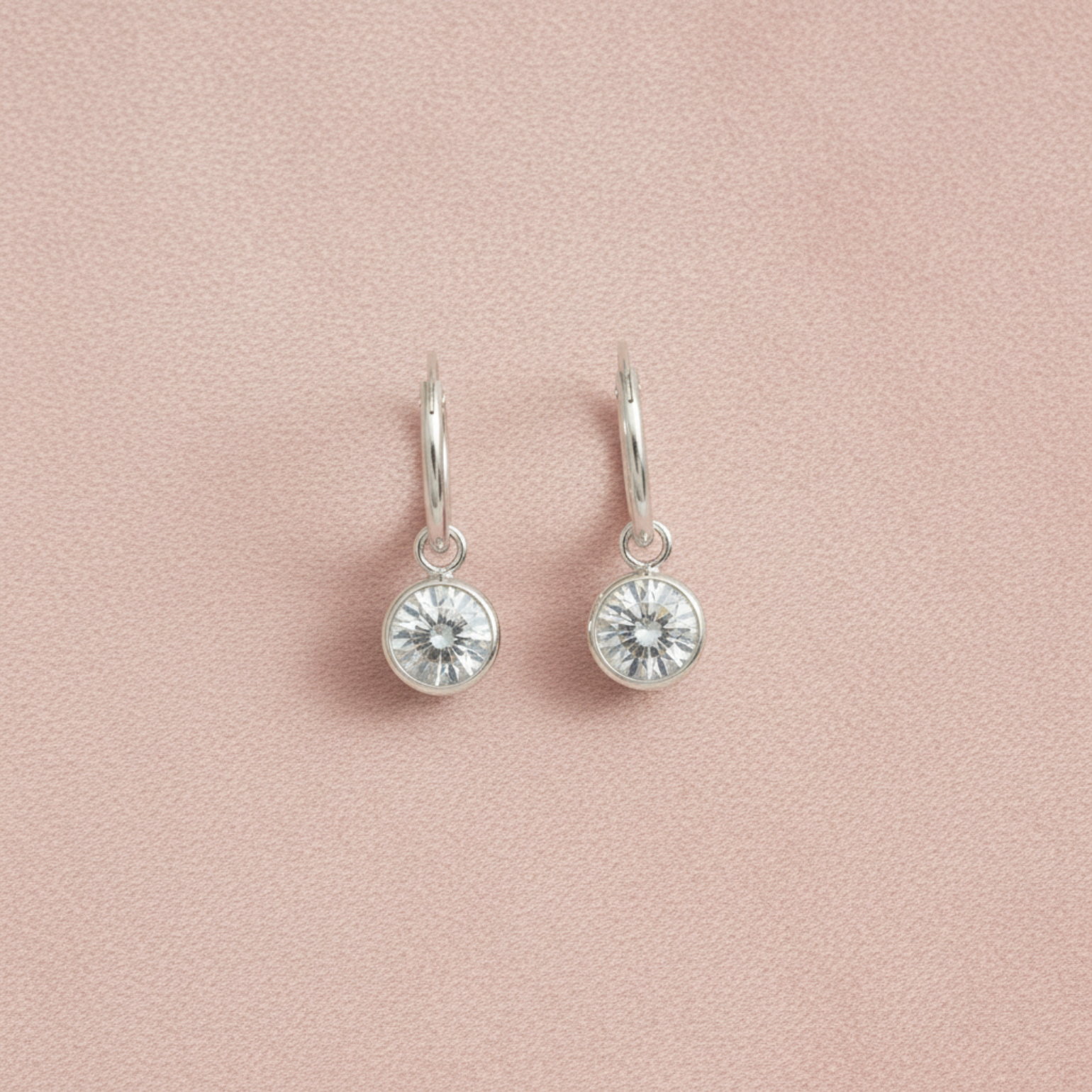 A pair of silver dangle earrings with round bezel-set clear crystals, displayed on a textured light pink background.