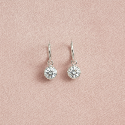 A pair of silver dangle earrings with round bezel-set clear crystals, displayed on a textured light pink background.