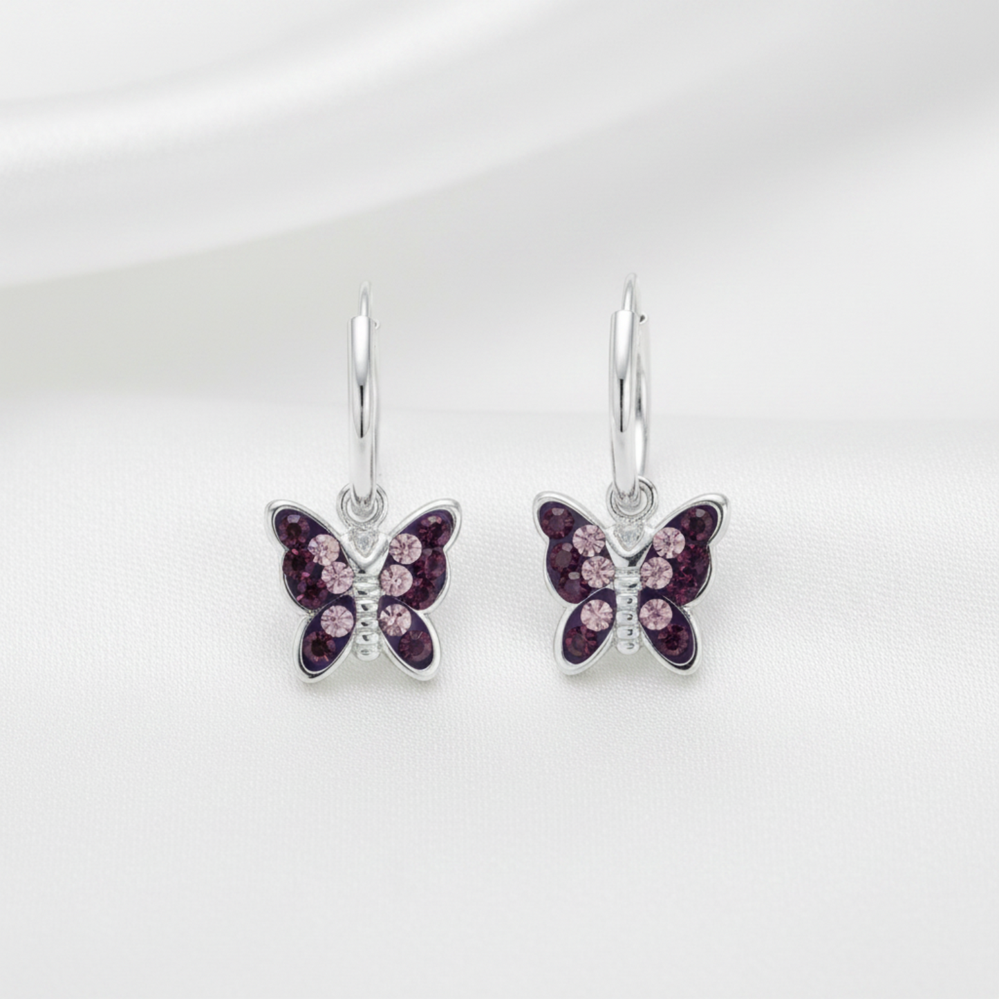 A pair of silver butterfly drop earrings with dark purple and light pink crystal details, displayed on a soft white fabric background.