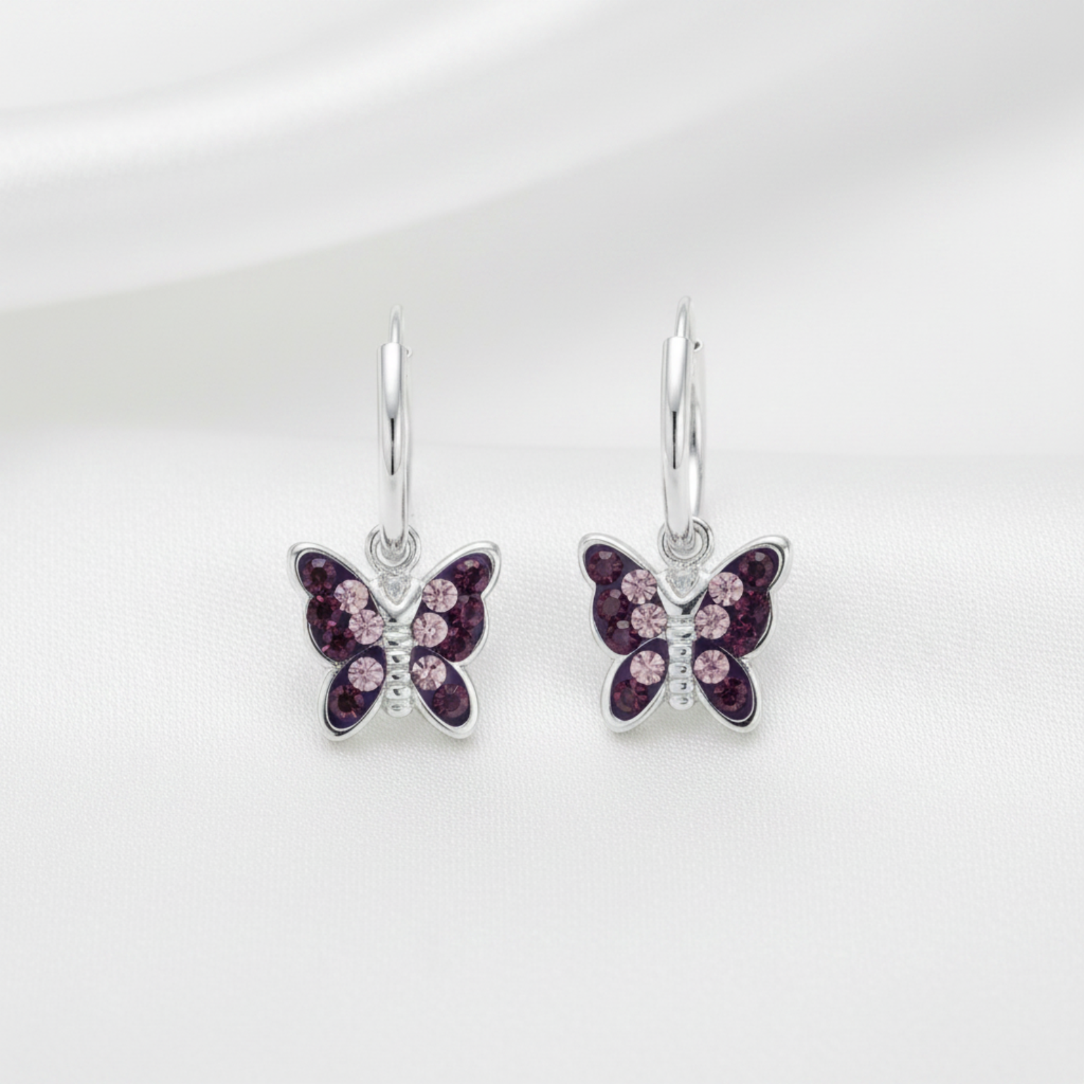A pair of silver butterfly drop earrings with dark purple and light pink crystal details, displayed on a soft white fabric background.