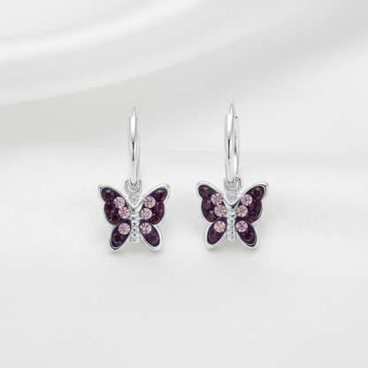 A pair of silver butterfly drop earrings with dark purple and light pink crystal details, displayed on a soft white fabric background.