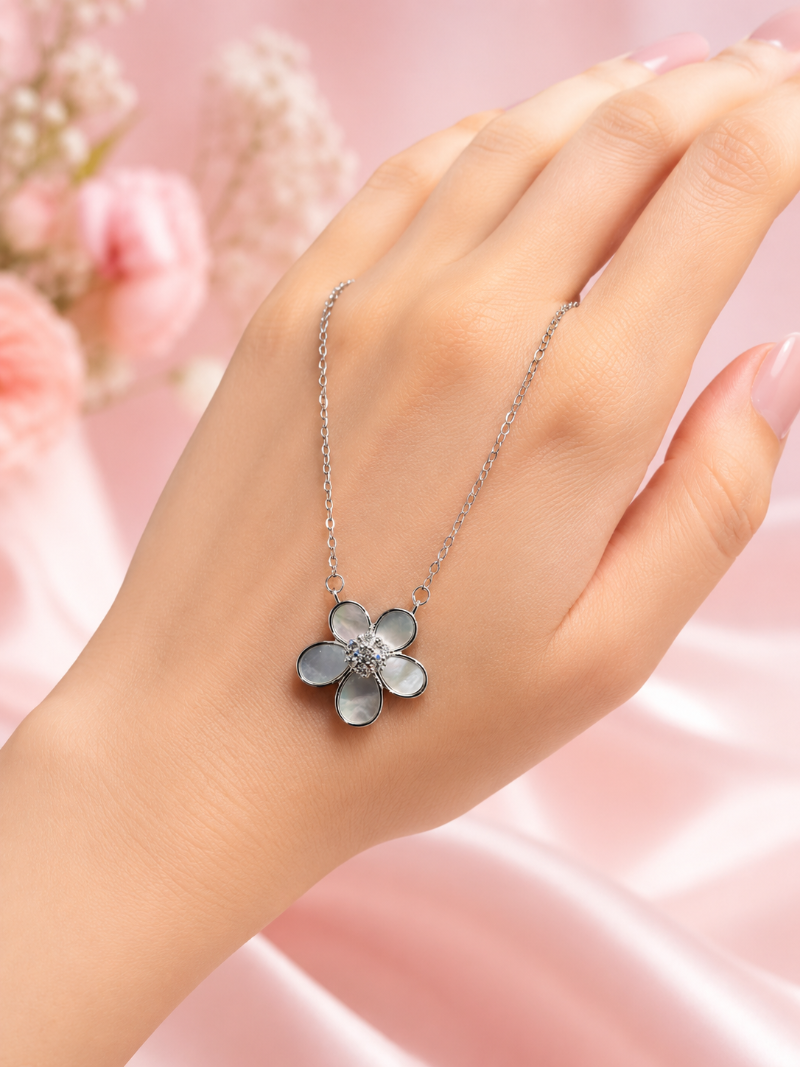 Hand wearing a silver necklace with a flower pendant against a soft pink background