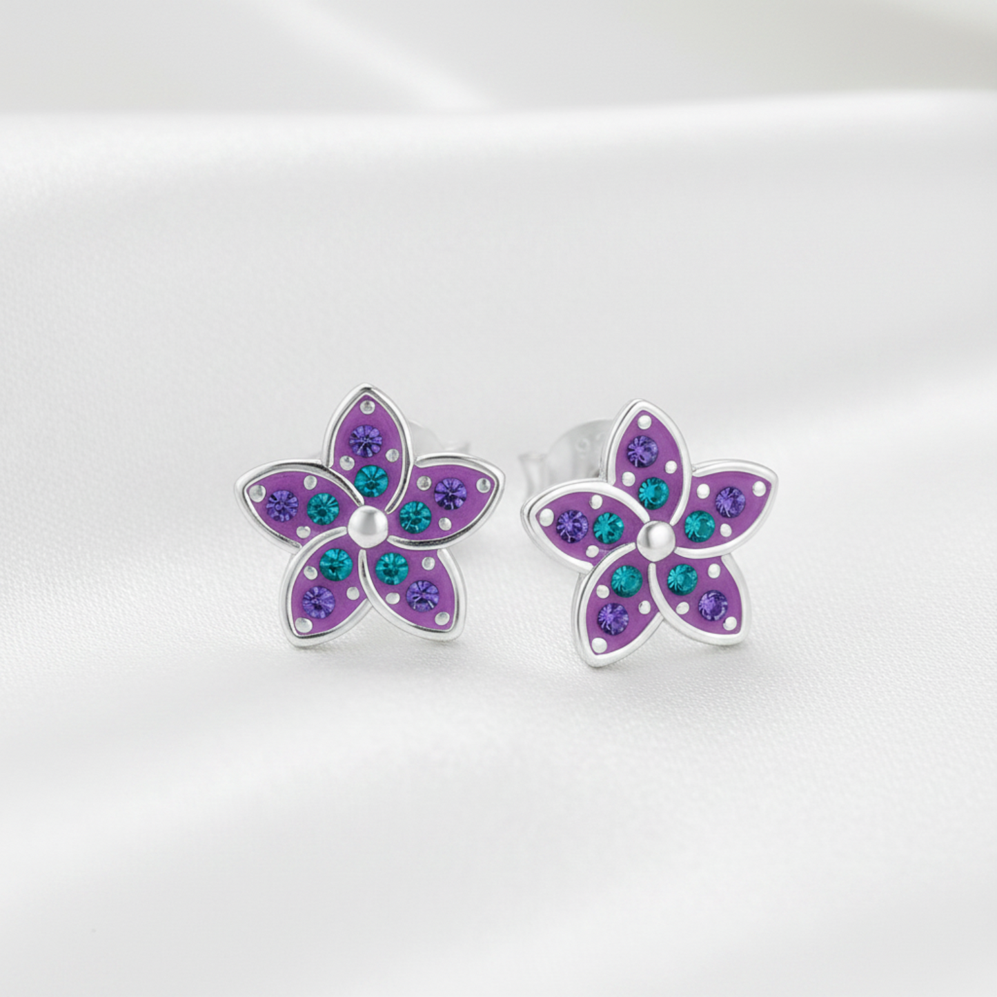 A pair of silver flower stud earrings with purple petals and small teal and purple rhinestones, displayed on a soft white satin background.