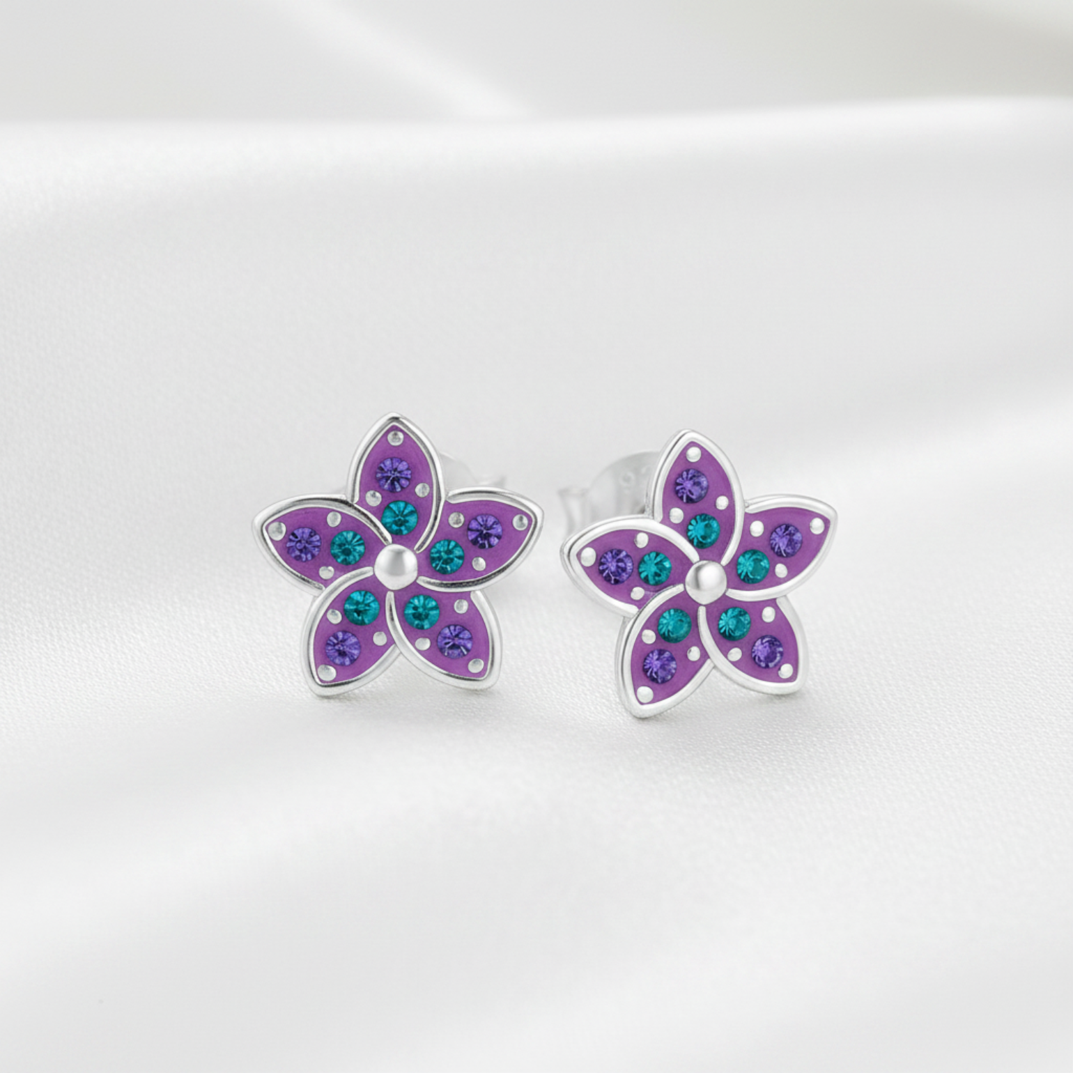 A pair of silver flower stud earrings with purple petals and small teal and purple rhinestones, displayed on a soft white satin background.
