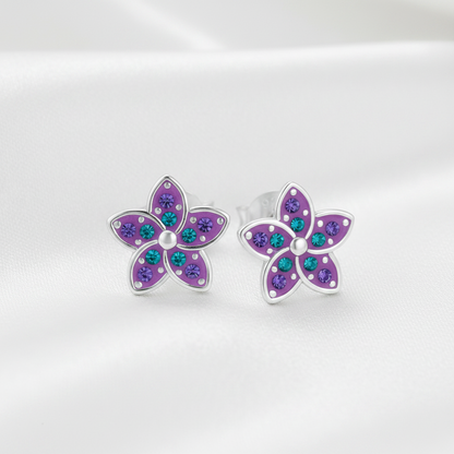 A pair of silver flower stud earrings with purple petals and small teal and purple rhinestones, displayed on a soft white satin background.