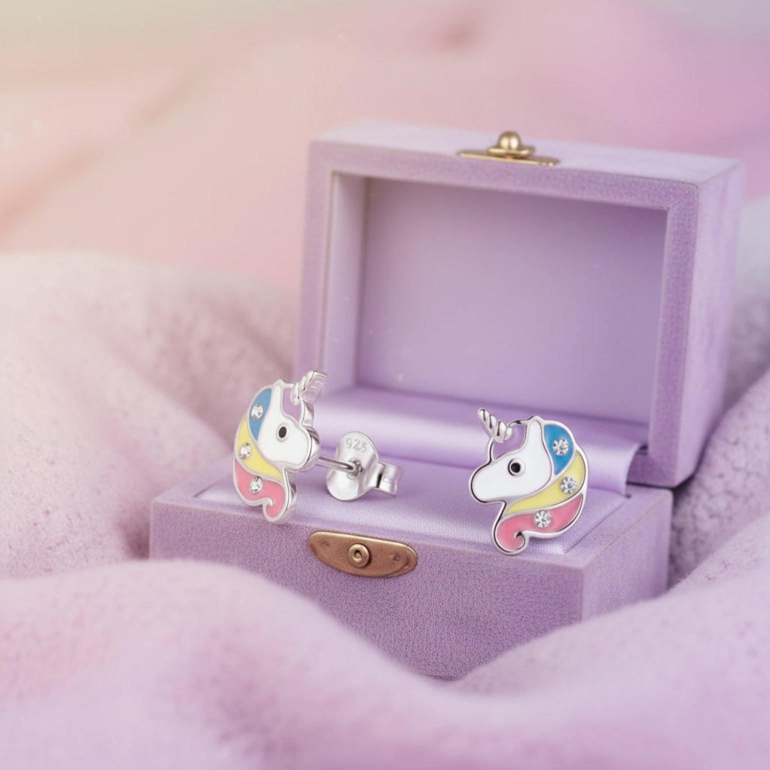 A pair of colorful unicorn stud earrings displayed in an open wooden jewelry box on soft gray fabric with a pink blurred background.