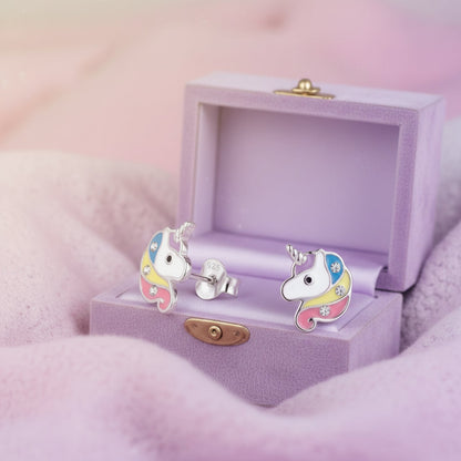 A pair of colorful unicorn stud earrings displayed in an open wooden jewelry box on soft gray fabric with a pink blurred background.