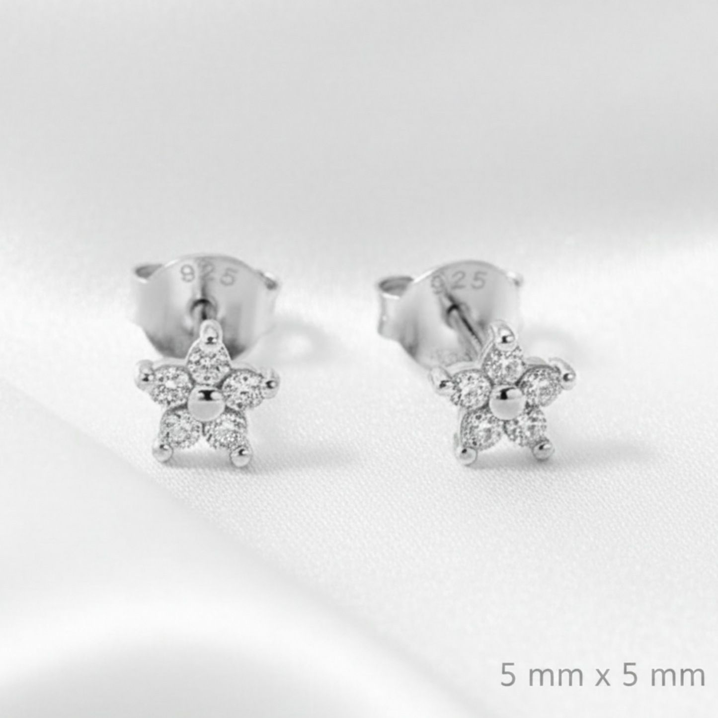 A pair of silver stud earrings with small star-shaped crystal clusters, displayed on a draped white satin fabric background.