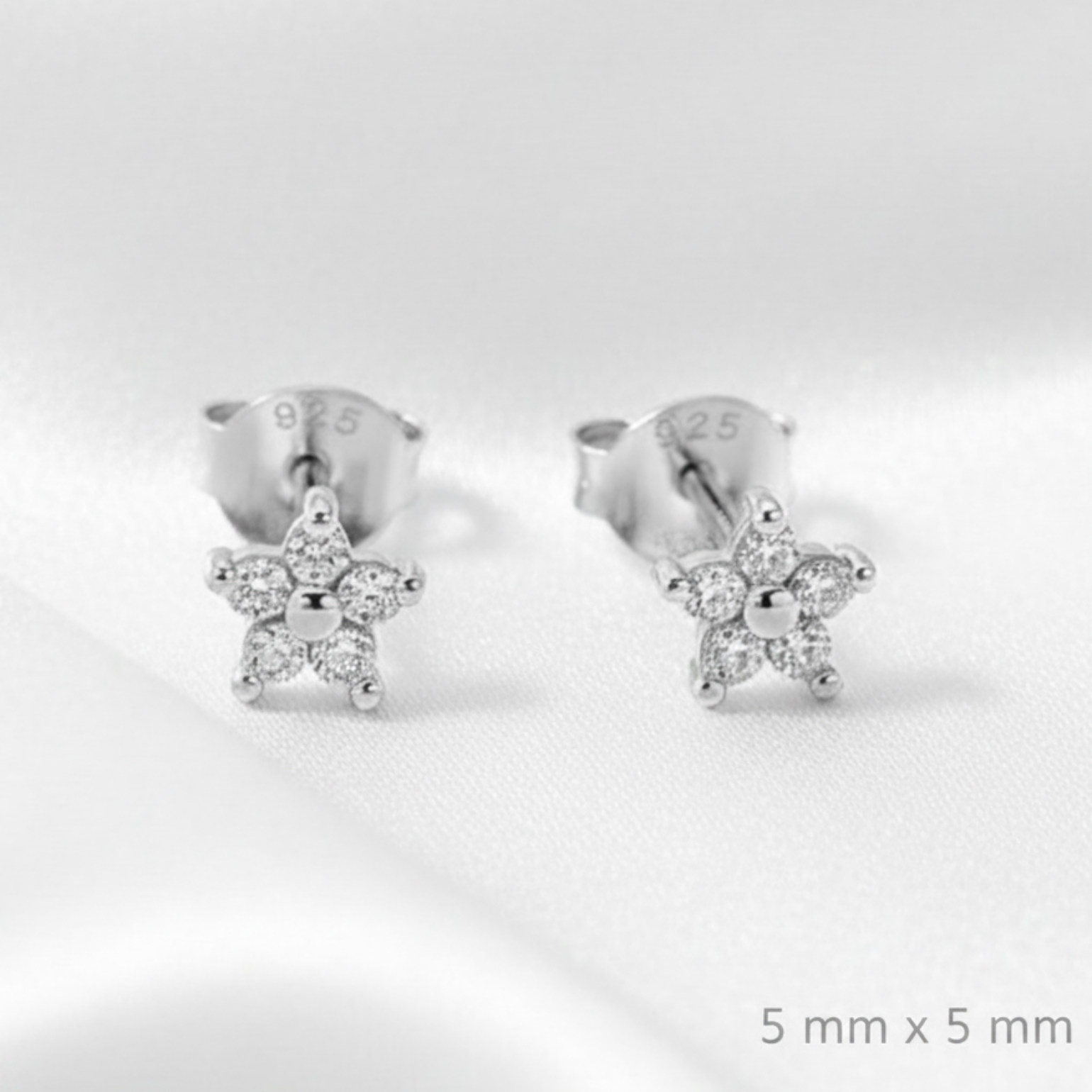 A pair of silver stud earrings with small star-shaped crystal clusters, displayed on a draped white satin fabric background.