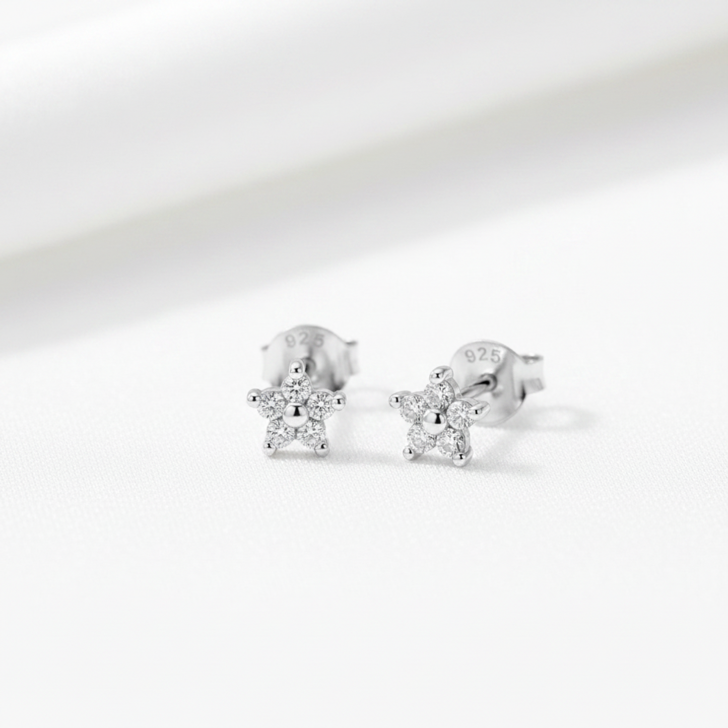A pair of silver star-shaped stud earrings with clear gemstones, displayed on a textured light green surface with white flowers and green leaves in the background.