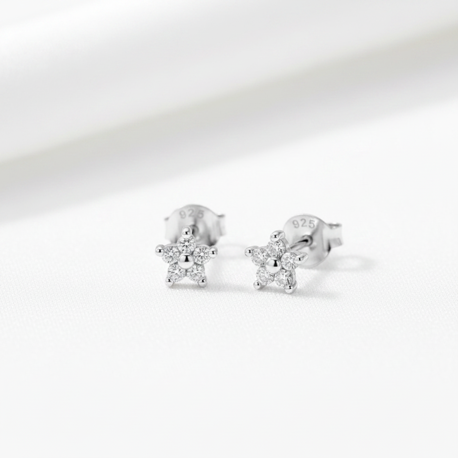 A pair of silver star-shaped stud earrings with clear gemstones, displayed on a textured light green surface with white flowers and green leaves in the background.