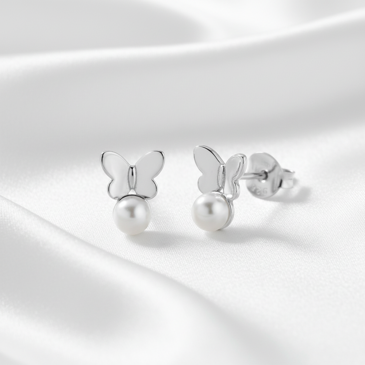 A pair of silver butterfly stud earrings with a dangling pearl, displayed on a soft, draped white satin fabric background.