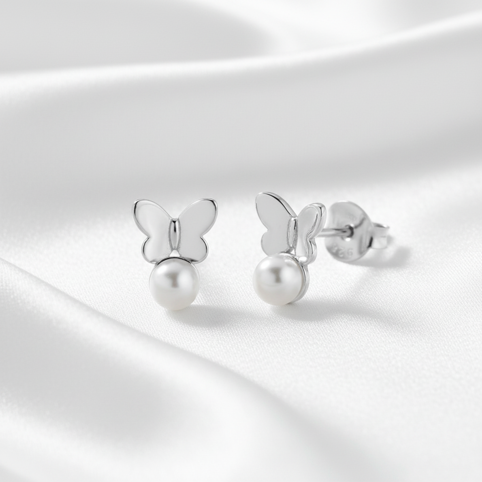 A pair of silver butterfly stud earrings with a dangling pearl, displayed on a soft, draped white satin fabric background.