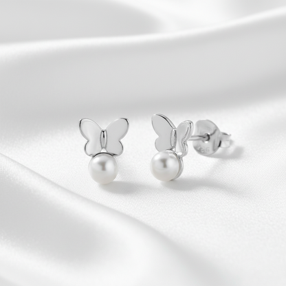 A pair of silver butterfly stud earrings with a dangling pearl, displayed on a soft, draped white satin fabric background.