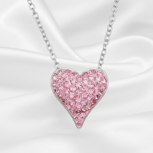 A silver heart pendant necklace paved with pink rhinestones, hanging from a silver chain against a draped white satin background.