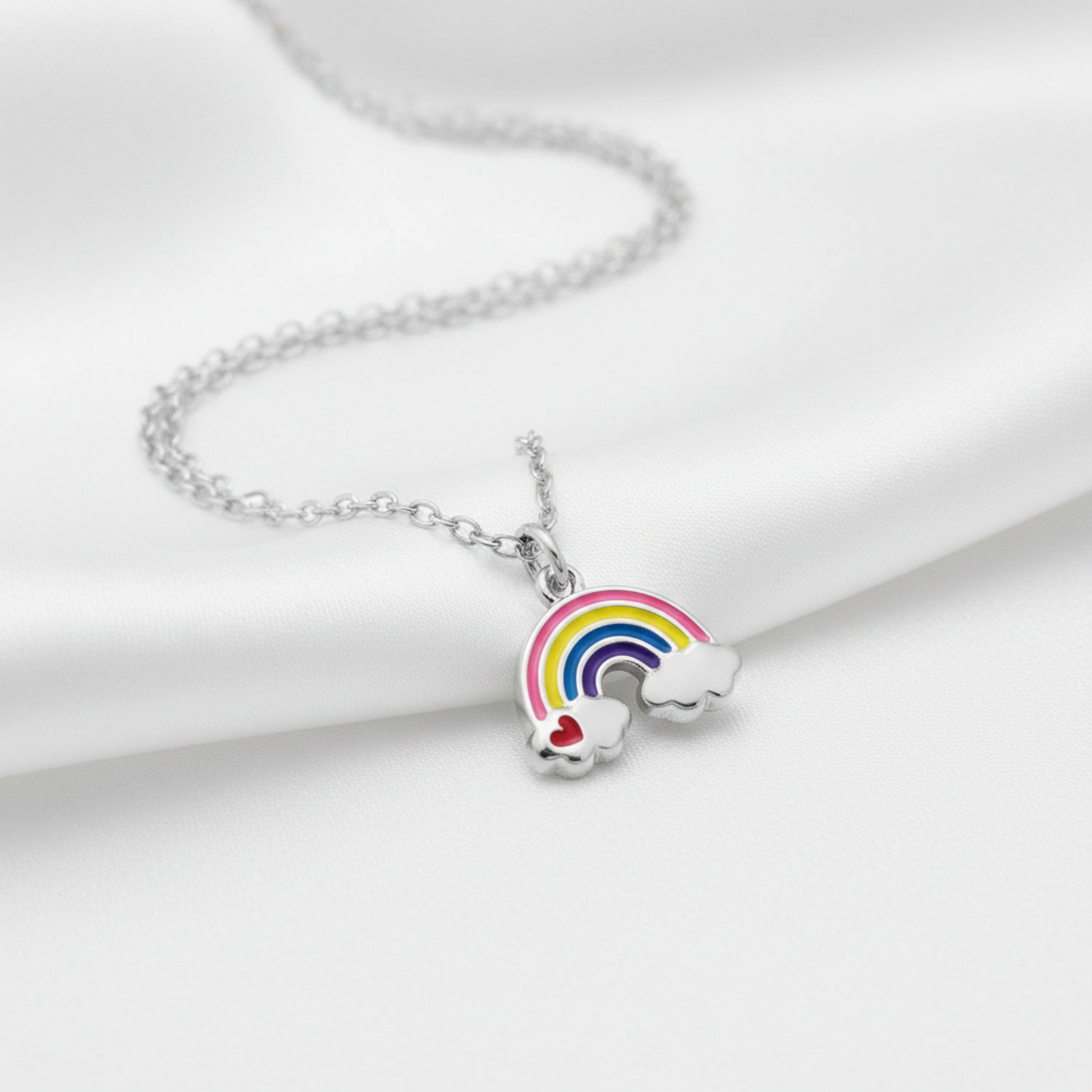 A silver chain necklace with a charm shaped like a rainbow arching over two small white clouds, one with a red heart, displayed on a soft white satin fabric background.