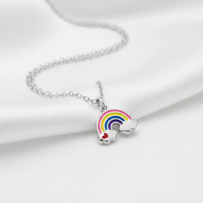 A silver chain necklace with a charm shaped like a rainbow arching over two small white clouds, one with a red heart, displayed on a soft white satin fabric background.