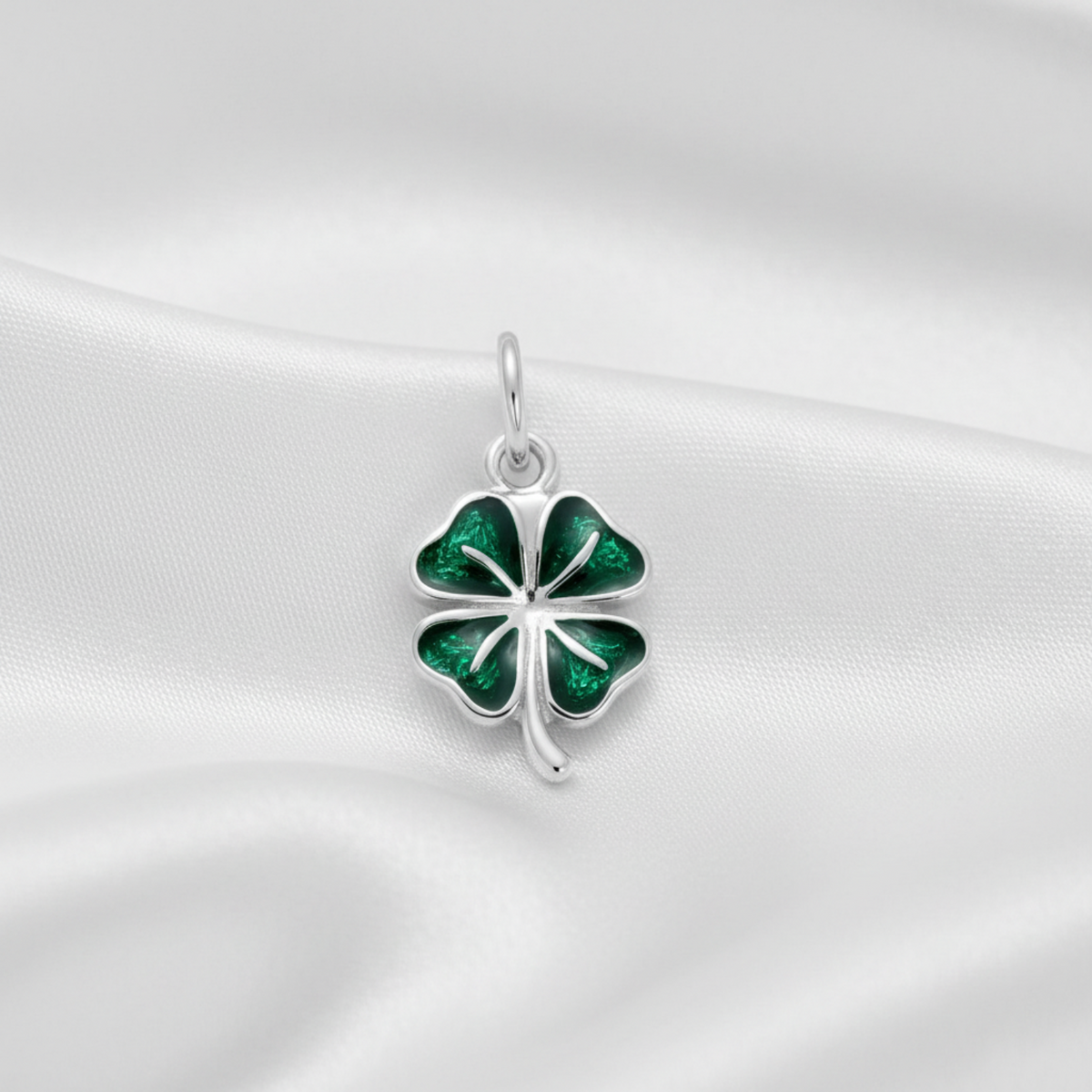 A sterling silver four-leaf clover charm with vibrant green enamel inlay, resting on a draped white satin fabric background.