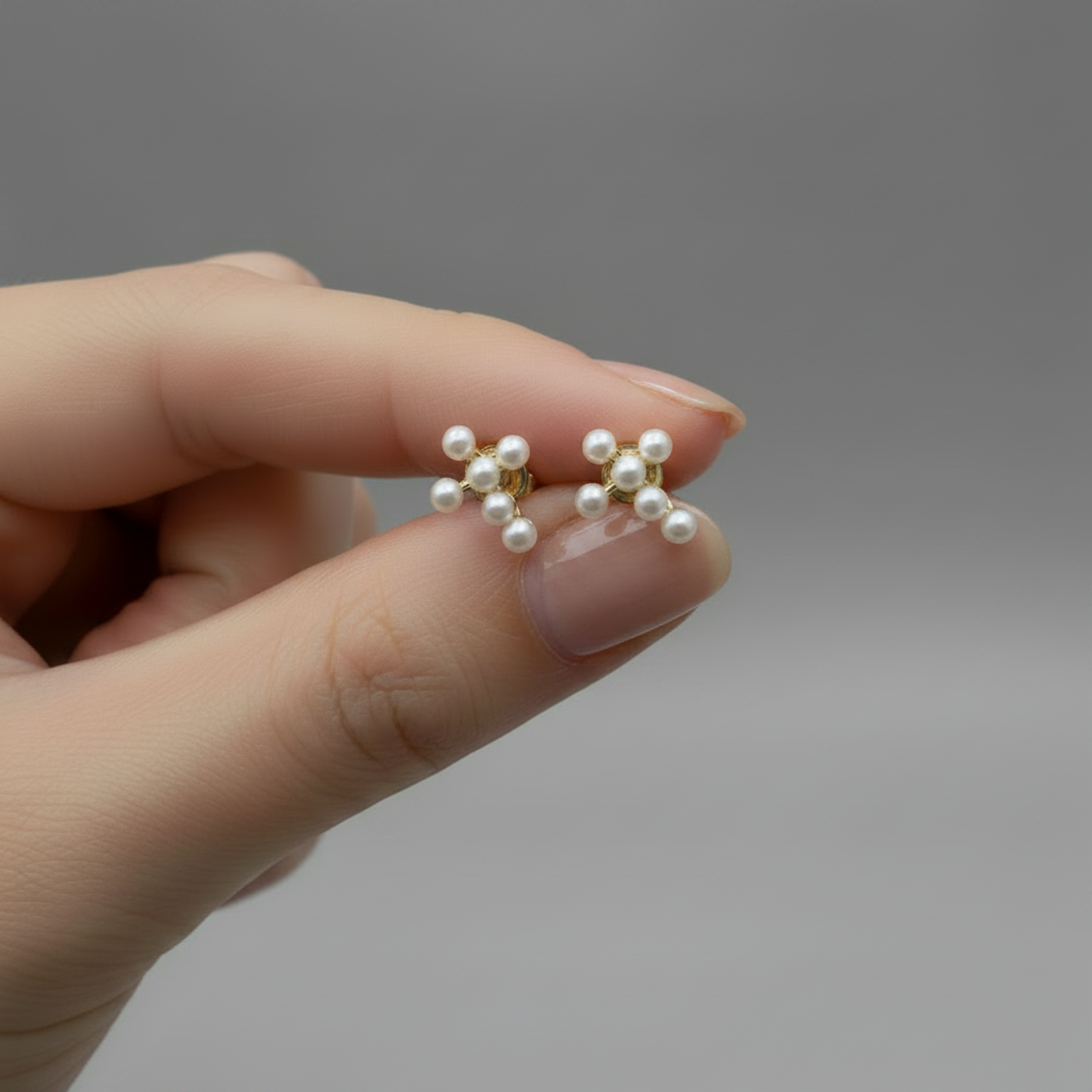 A close-up of a hand holding a pair of small gold stud earrings with clustered white pearls against a soft gray background.