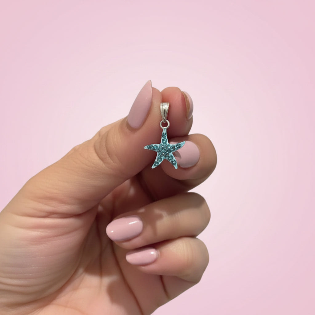 A hand with light pink manicured nails holds a small silver starfish pendant embellished with light blue rhinestones against a blurred background.