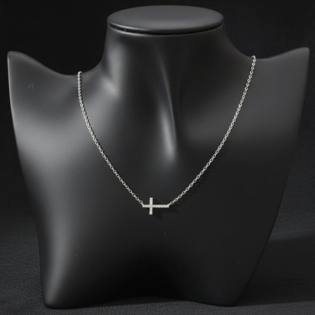 A delicate silver cross pendant necklace displayed on a matte black jewelry bust against a light gray background.