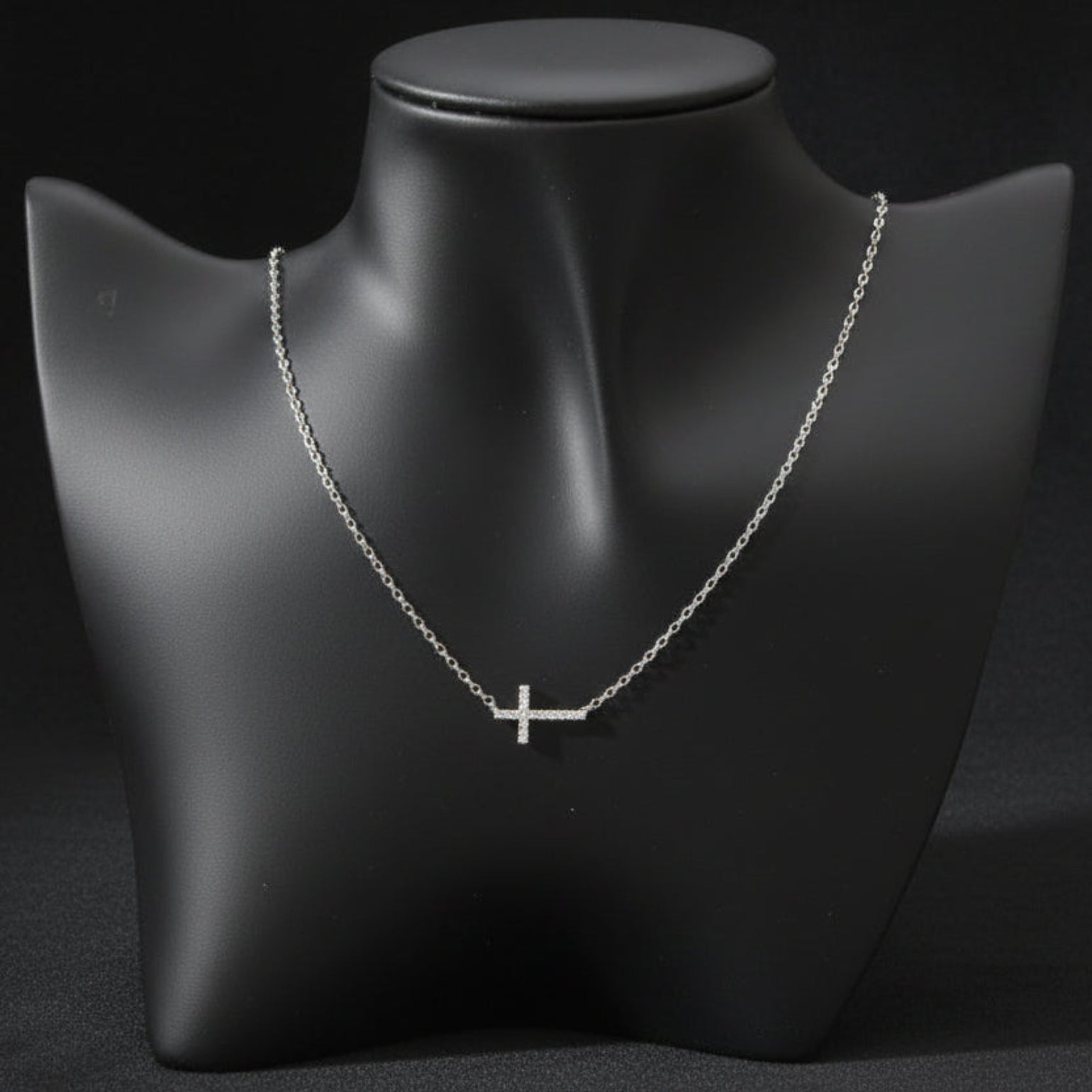 A delicate silver cross pendant necklace displayed on a matte black jewelry bust against a light gray background.