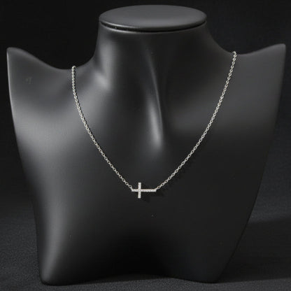 A delicate silver cross pendant necklace displayed on a matte black jewelry bust against a light gray background.