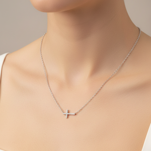 A close-up of a model wearing a delicate silver cross pendant necklace with small red gemstone accents on the ends, styled with a white top against a soft beige background.