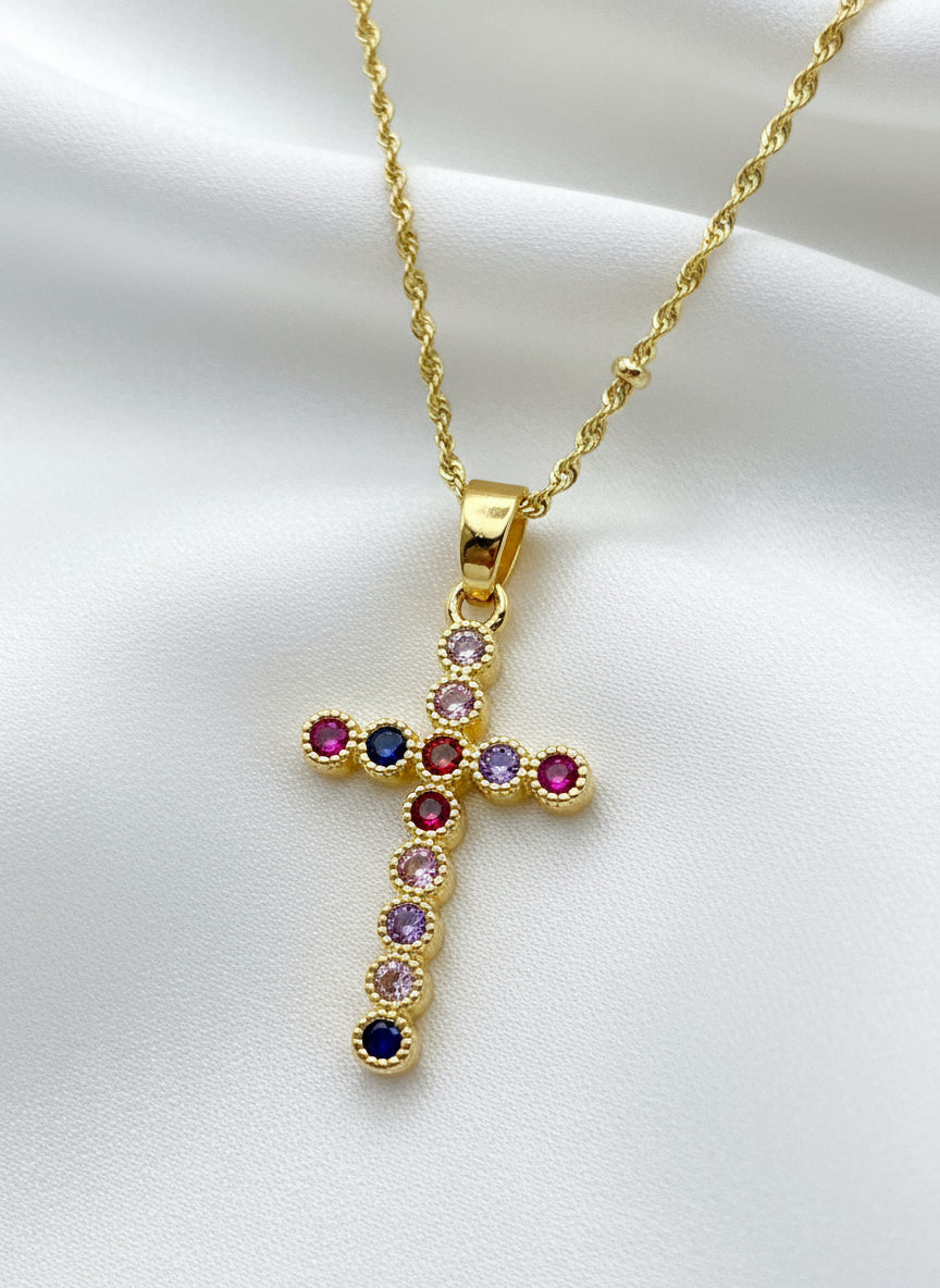 Gold cross pendant with colored gemstones on a light pink background