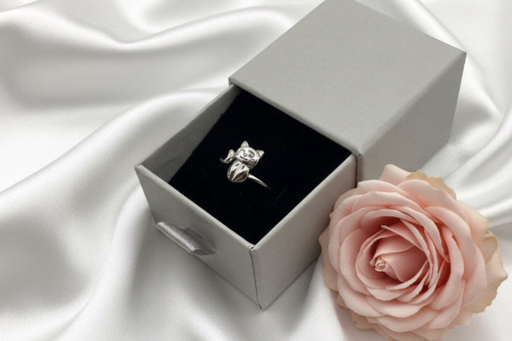 Silver ring in a white box with a pink rose on a white fabric background