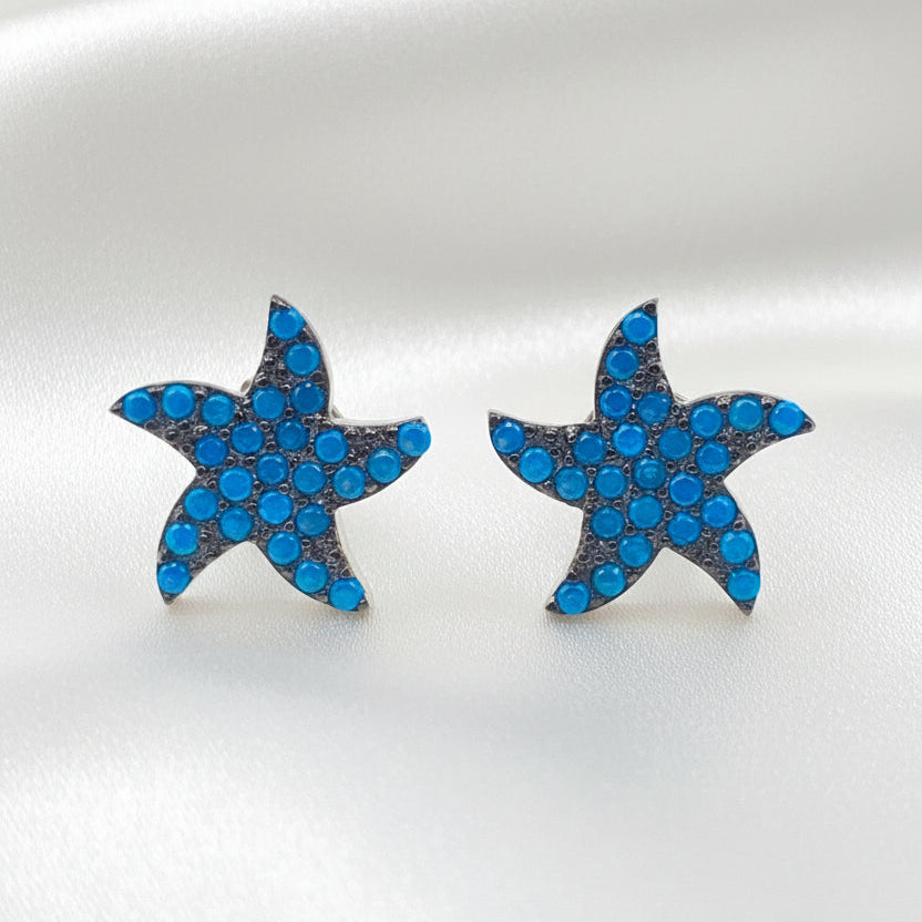 A pair of starfish-shaped stud earrings covered in small blue rhinestones with silver-toned edges, displayed on a plain white background.