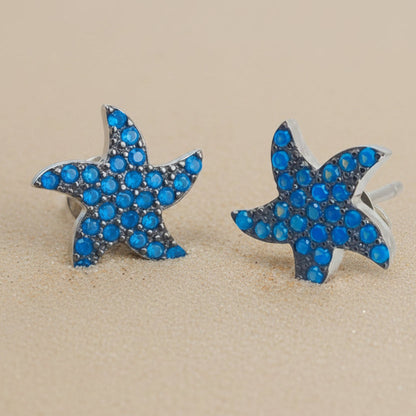 A pair of starfish-shaped stud earrings with bright blue rhinestones set in silver-tone metal, displayed on sand.
